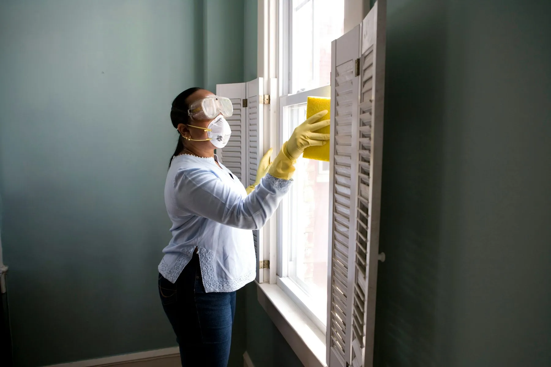 A cleaner in goggles, a mask and yellow gloves wiping a window frame with a sponge